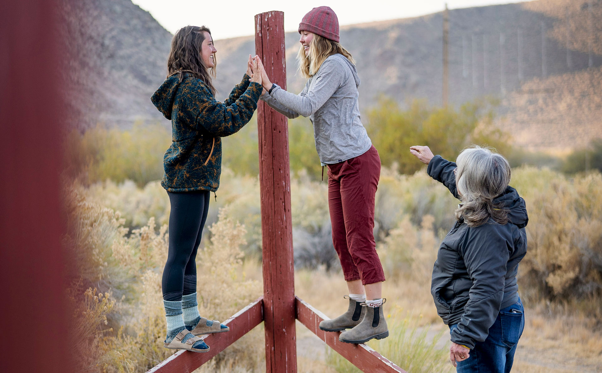 Two women practice balance on a wooden outdoor structure while an instructor guides them, symbolizing peer support.