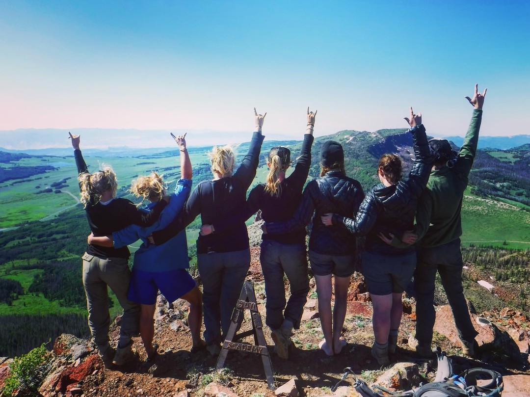 A group stands arm-in-arm on a mountain peak, celebrating their shared accomplishment and symbolizing the empowerment, confidence, and resilience gained by overcoming challenges in wilderness therapy.