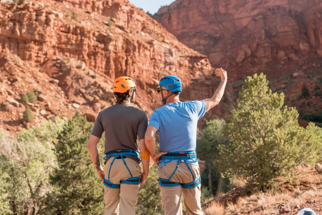 A mentor gestures toward the red rock canyon ahead, encouraging a young man during a men's adventure therapy program focused on mental health and substance abuse recovery.