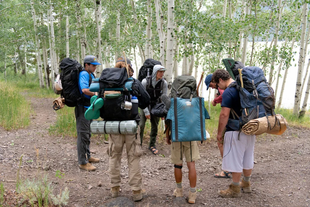 A group of young men huddle with their backpacks on, engaging in group processing before a wilderness expedition—a therapeutic approach that builds peer support, communication, and resilience.