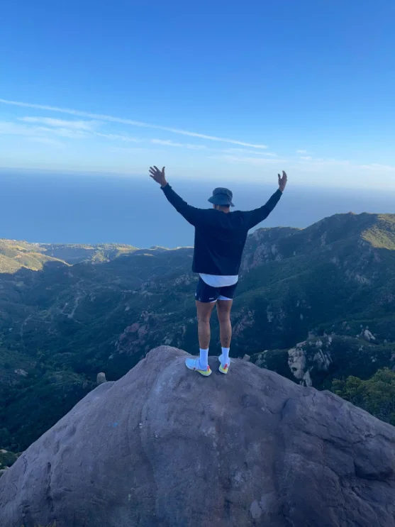 A young man stands triumphantly on a mountain peak with his arms raised, overlooking the ocean, symbolizing the freedom, hope, and personal empowerment achieved through an adventure therapy program for mental health.