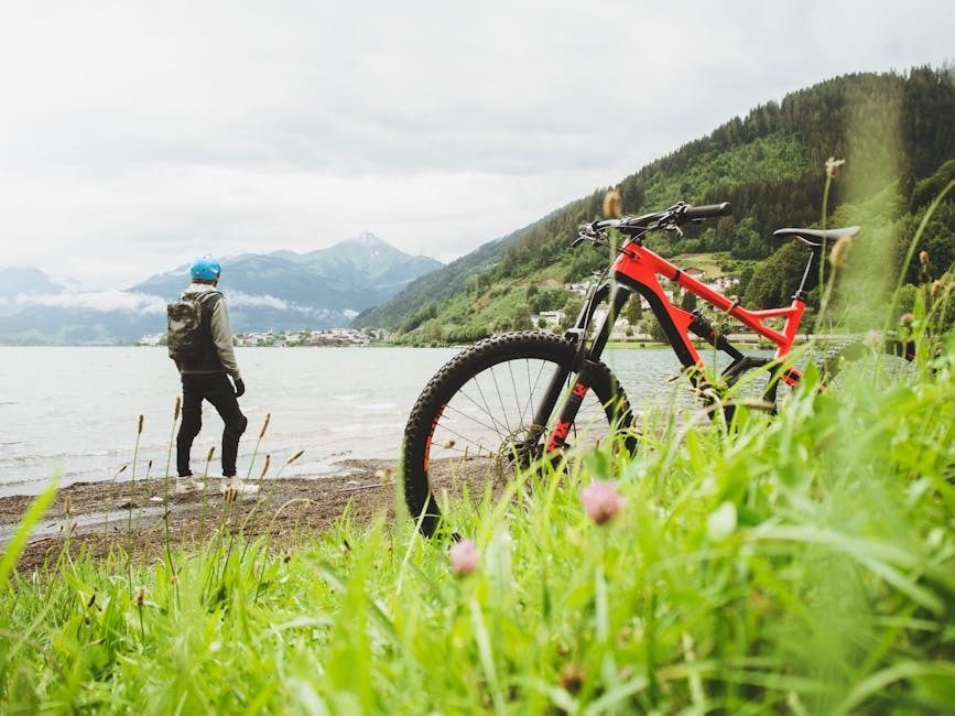 A person wearing a helmet stands on a lake shore looking at mountains, next to a red mountain bike.